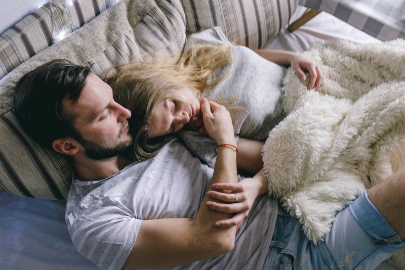 A couple cuddle together in bed, dressed in day clothes and with blankets.
