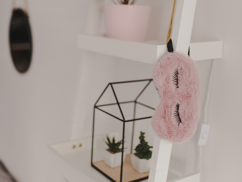 A pink mask with eyelashes hands on a bookshelf in a white room with two succulent plants behind it.