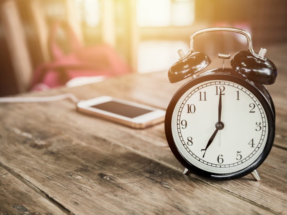 A phone sits on a wooden table, plugged in to charge. Next to it is an old-fashioned alarm clock.