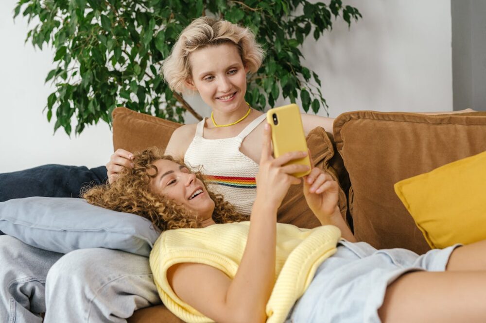 Two women on a couch: one sitting upright and the other resting with her head on the lap of the upright woman.