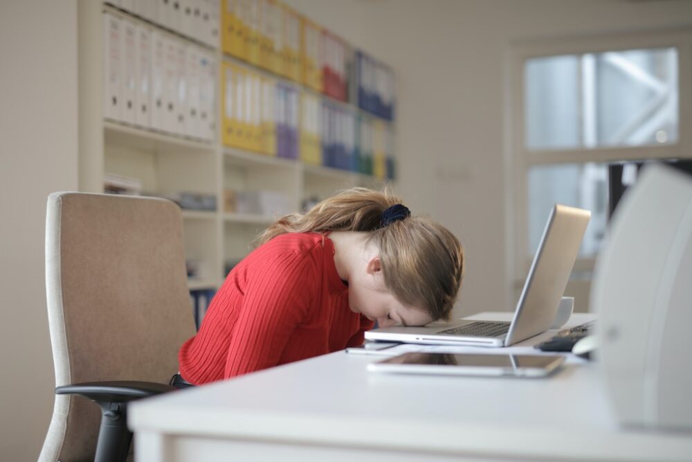 Morning Grogginess: Waking Up Tired With Sleep Inertia - A woman in a red shirt sleeping on her laptop at work.