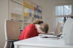 Morning Grogginess: Waking Up Tired With Sleep Inertia - A woman in a red shirt sleeping on her laptop at work.