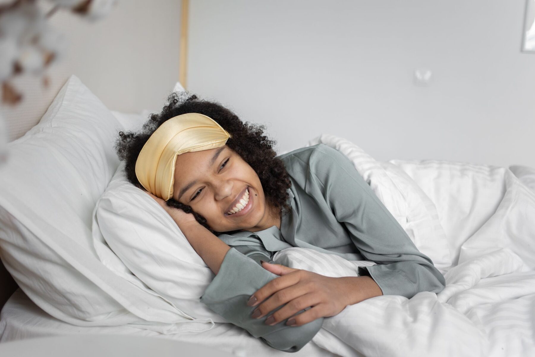 A joyful woman relaxing in a cosy bedroom, wearing a satin sleep mask and smiling brightly.