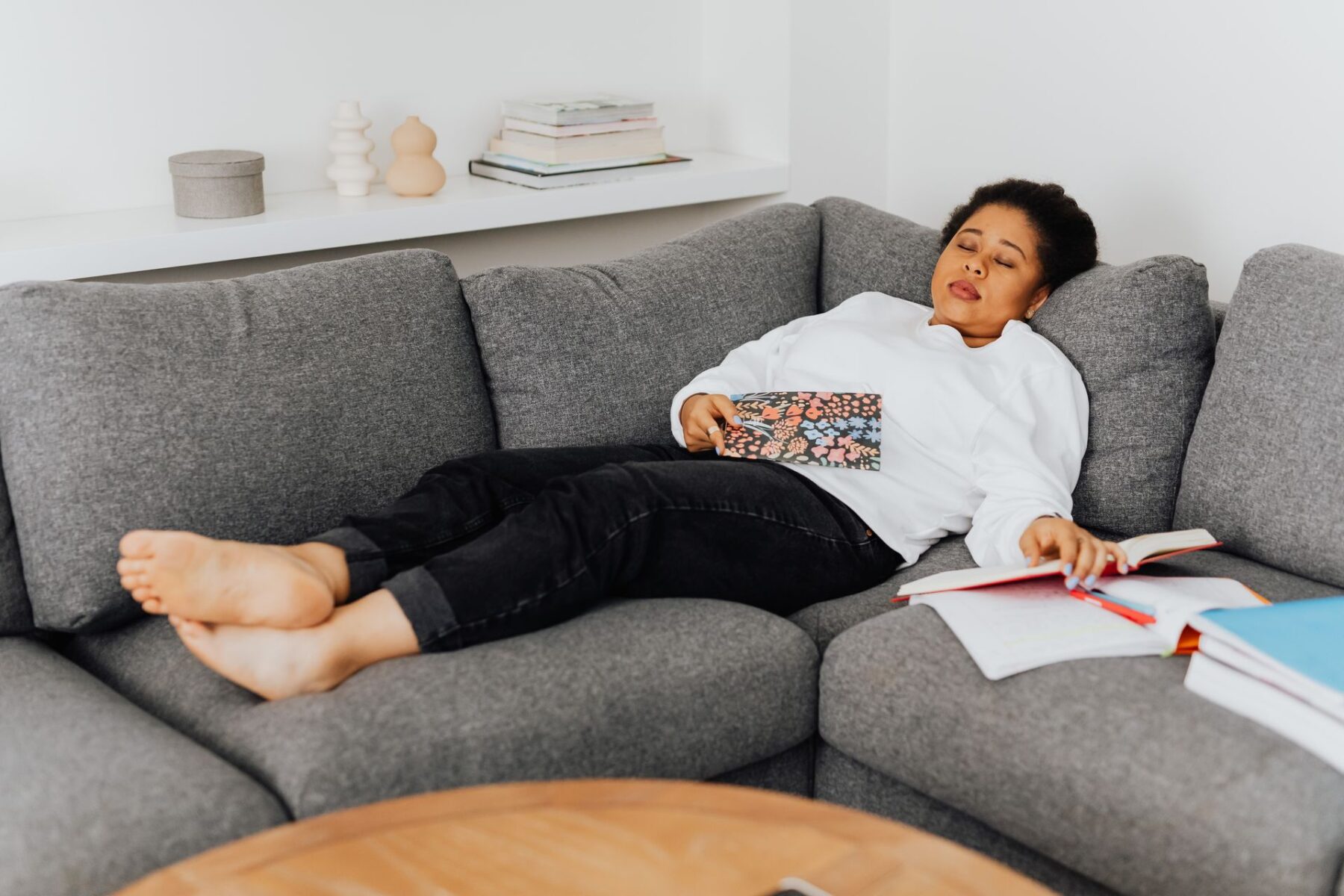 A young woman resting on a cozy grey sofa with open books around her, appearing tired after studying.