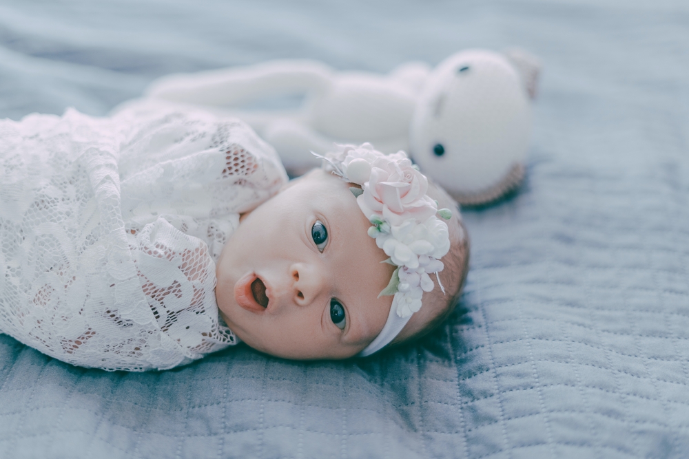 A baby swaddled in a white blanket, with a white headband, on a grey bed next to a white soft toy
