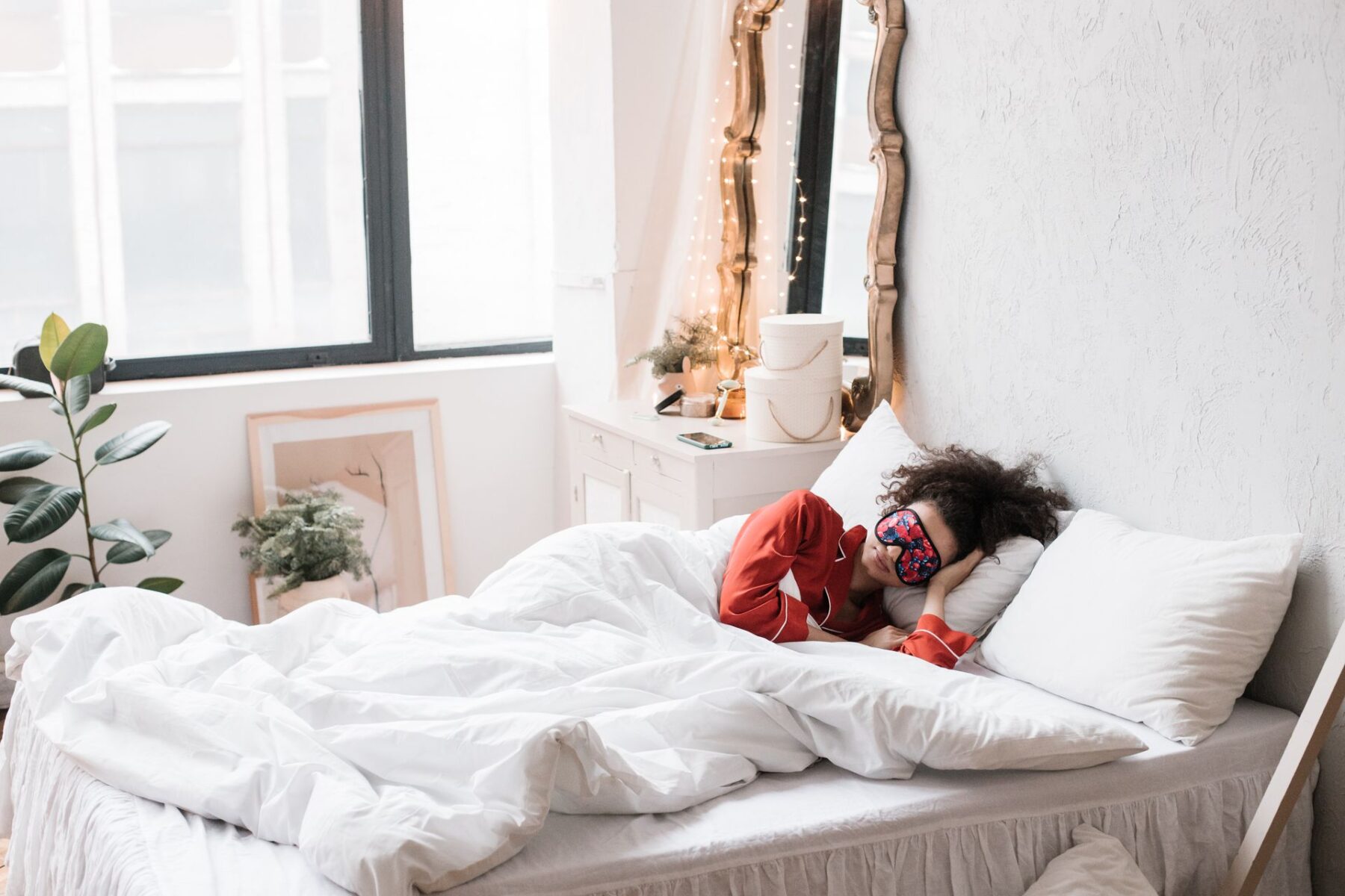 Woman wearing a sleep mask, resting on a cosy white bed surrounded by indoor plants in a bright bedroom.