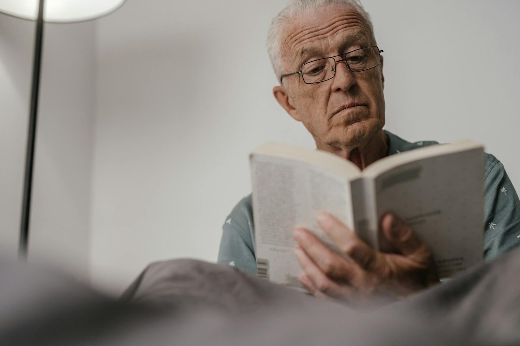 Elderly man with glasses reading book indoors, focused expression.