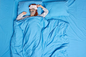 woman-lies-comfortable-bed-blanket-applies-facial-mask-wears-sleepmask-pajama